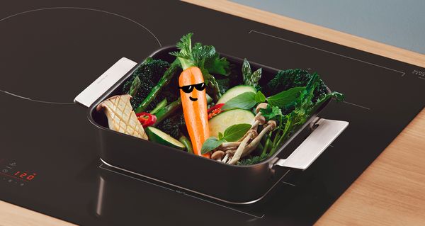 Induksjonsplatetopp i Bosch Serie 6 med en stekepanne med grønnsaker og en gulrot med solbriller.