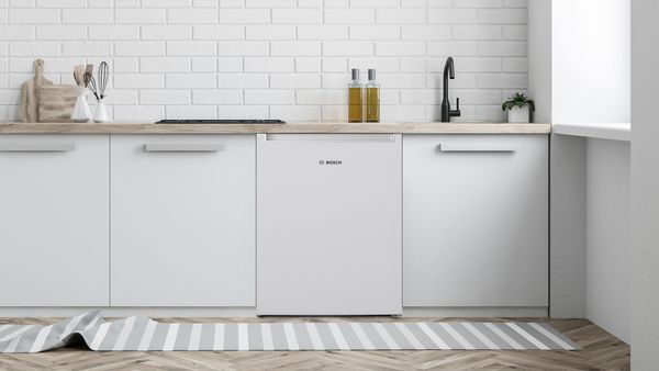 Et mini-køleskab er anbragt imellem hvide skabe under et køkkenbord.