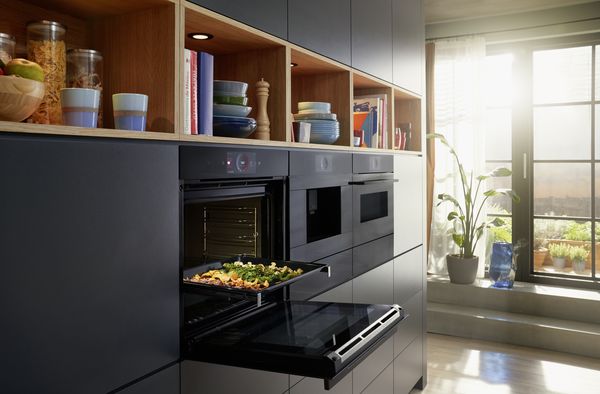 Cuisine élégante avec four intégrable Série 8 de Bosch avec fonction Air Fry et tiroir culinaire chauffant. Cuisson de différents légumes au four. Plus de légumes sur une planche à découper déposée sur un comptoir.