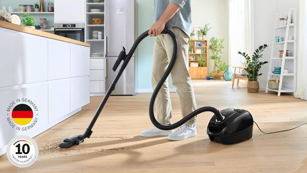 A man uses a cylinder vacuum to clean the floors near a kitchen island in a warm, bright home.