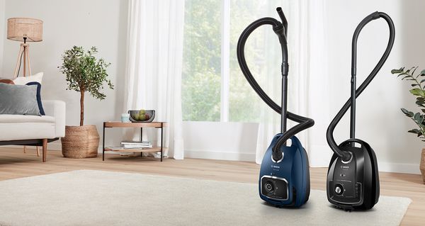 A pair of Bosch cylinder vacuums are parked on the edge of a neutral-coloured carpet in a light-flooded living room.