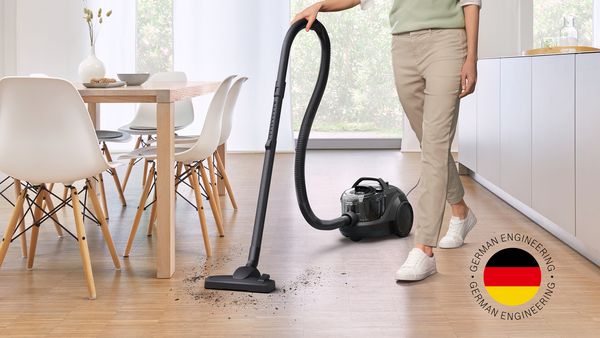 A woman uses a cylinder vacuum to clean the floors around a small kitchen table and chairs next to a kitchen island in a warm, bright home.