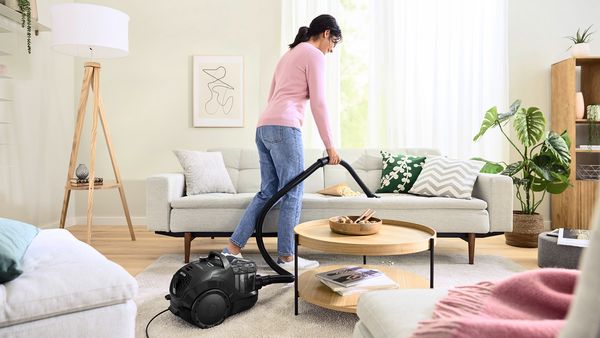 A woman uses a cylinder vacuum to clean the couch upholstery in a warm, bright living room.