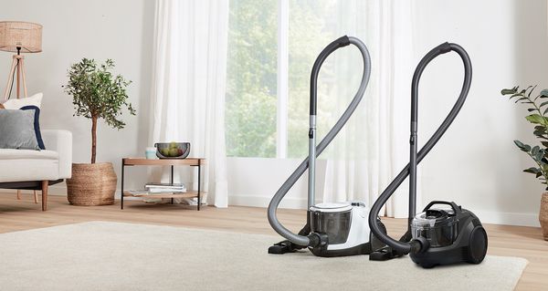 A pair of cylinder vacuums stand on the edge of a light-coloured carpet in a warm, bright living room.