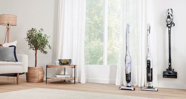 Three cordless vacuums, two standing upright and one mounted on the wall, are positioned alongside the large window of a warm, bright living area.