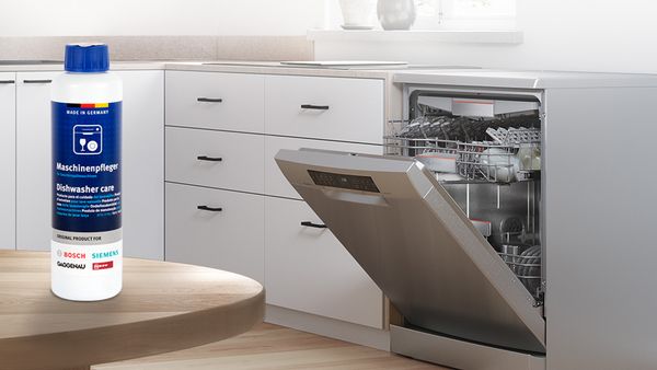 Продукт за почистване на съдомиялна машина стои на дървена маса в светла бяла кухня с открехната съдомиялна машина Bosch на заден план.
