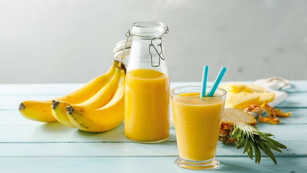 Banana smoothie in a glass next to a glass bottle with same smoothie and bananas, and sliced pineapple and mango.