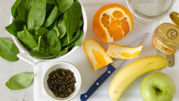 Orange, apple and banana next to a bowl of fresh spinach, seeds and honey shot from above.