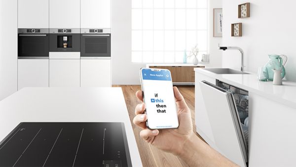 Mobilni uređaj koji se drži ispred kuhinje i snima otvorenu mašinu za pranje sudova. Na ekranu telefona prikazana je poruka „if this then that“ (ako se to dogodi, izvršiće se sledeće), unoseći atmosferu moderne pogodnosti.