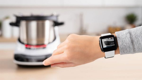 I forgrunden ses en hånd med et stilfuldt smartwatch omkring håndleddet. I baggrunden ses et Bosch Cookit produkt.