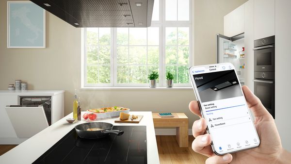 Ruka držiaca smartfón, na ktorom sa zobrazuje nastavenie odsávača pár. V pozadí na sporáku leží panvica, ktorá dotvára atmosféru kuchyne.