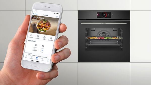 Smartphone che visualizza una ricetta di chips di verdure, tenuto di fronte a un forno che sta cuocendo proprio quel piatto. Il funzionamento del forno è sapientemente gestito dal telefono, per un'esperienza di cottura ideale.