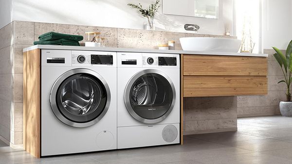 Duas máquinas de lavar roupa colocadas ao lado de um lavatório de casa de banho. No topo da máquina de lavar roupa à esquerda encontram-se duas toalhas verdes.