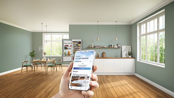 En smartphone viser Home Connect-startskærmen med en køkkenenhed og et åbent køleskab/fryser i baggrunden.