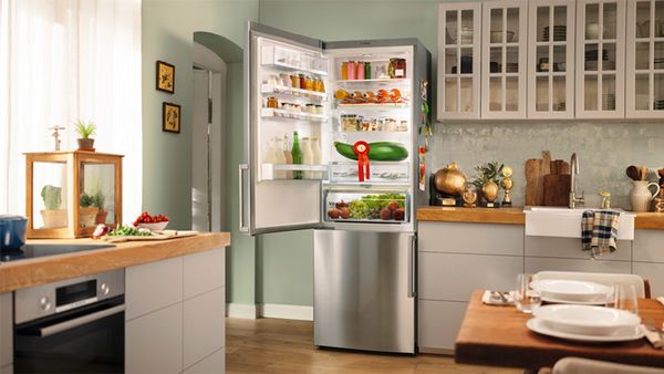 Eine Küche mit einem Arbeitstisch links und einem Esstisch rechts. Im Mittelpunkt steht ein gut gefüllter Kühlschrank mit einer geöffneten Tür, die den Blick auf eine Reihe von Lebensmitteln freigibt.