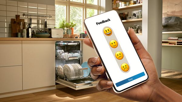 En smartphone viser Home Connect-appen og beder om feedback efter afslutning af en opvaskecyklus. Der er forskellige emojis at vælge imellem. I baggrunden ses en åben opvaskemaskine.