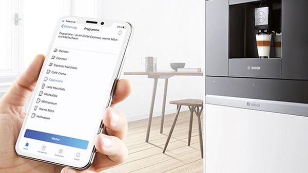 Uma mão segura um telemóvel que mostra a aplicação Home Connect com o modo que apresenta as definições para um cappuccino. Ao fundo, vamos uma máquina de café encastrada na parede.