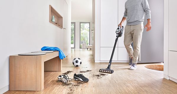 En mann støvsuger gulvet i en hall med en Unlimited 8 trådløs støvsuger og fjerner smuss ved siden av fotballsko og en benk.