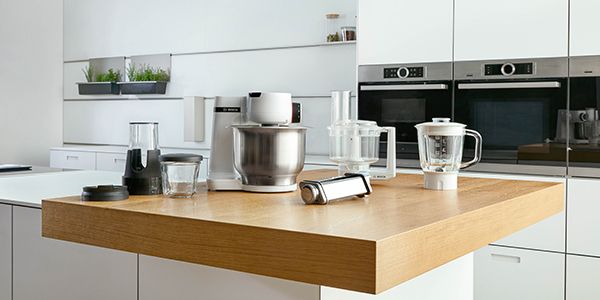 Inmitten verschiedener frischer Lebensmittel befindet sich eine Bosch Küchenmaschine in Weiß mit Edelstahlschüssel.