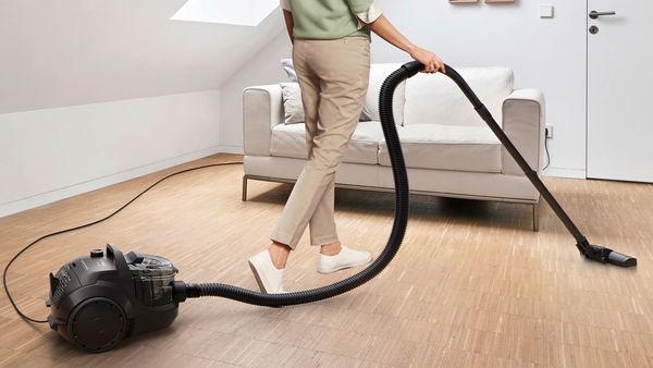 Eine Frau saugt mit einem Staubsauger ohne Beutel von Bosch der Serie 4 in einem modernen Wohnzimmer unter dem Dach einen Hartboden.
