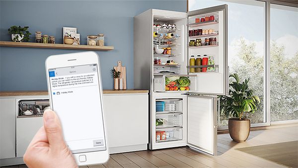 Mâini ținând un smartphone într-o bucătărie modernă. Pe ecranul telefonului este afișată aplicația Home Connect. Este vizibilă o combină frigorifică cu ușile deschise.