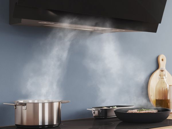Dampf wird von einem Topf und einer Bratpfanne auf einem Kochfeld in Richtung einer an der Wand montierten Dunstabzugshaube gesaugt.
