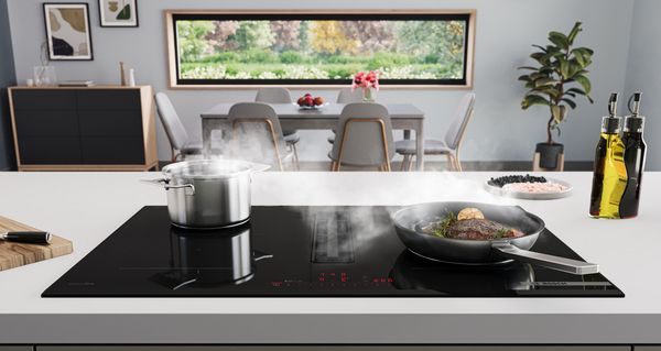 Bosch-kookplaat met geïntegreerde ventilatiemodule. Een braadpan met een biefstuk en een kookpan op de kookplaat, eetruimte met eettafel op de achtergrond.