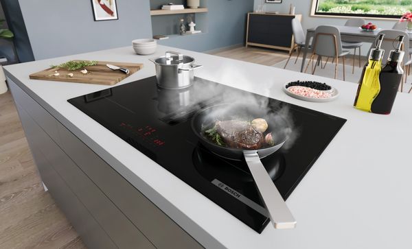 Schuin perspectief van de Bosch kookplaat met geïntegreerde afzuigmodule met een biefstuk die in een pan wordt gebakken en groenten die in een pan koken.