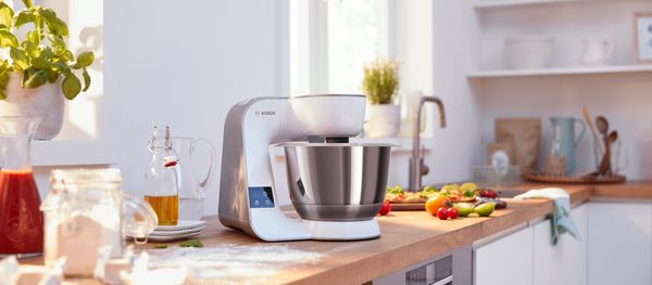 Optimum MUM Series 8, hand blender ErgoMaster Series 6, blender VitaPower Series 4, on a wooden countertop. Several people operate the devices. Knead dough, roll out dough. Mixing ingredients. Flour, sugar, fruits, nuts are on the table.