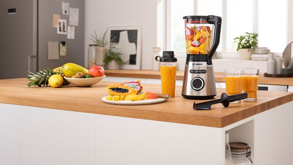 Exotische Früchte werden im VitaPower Serie 4 von Bosch auf einer modernen Kücheninsel zu einem Smoothie gemixt. Daneben stehen zwei frische Smoothies, Schalen mit Obst und ein Smoothie in einem ToGo-Becher.