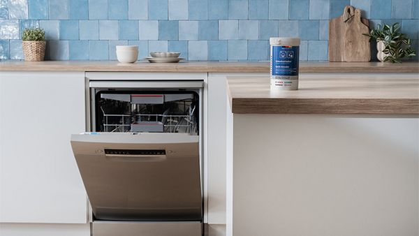 W kuchni o jasnych kolorach zainstalowana jest zmywarka ze stali nierdzewnej z otwartymi drzwiczkami i butelką produktu do pielęgnacji stojącą na drewnianym blacie.