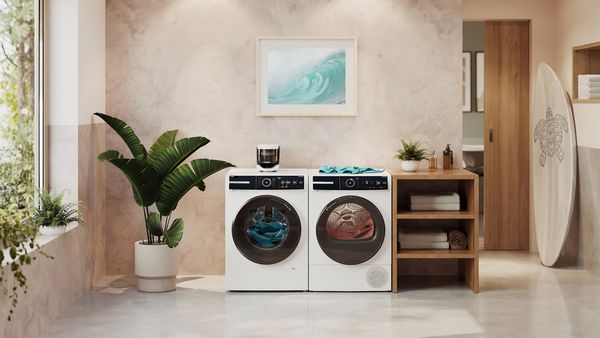 A washing machine with a Bosch Microplastic Filter and a dryer stand below a framed image of a wave in a bright laundry room filled with plants.