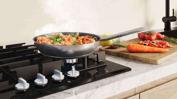 Poêle avec des pâtes aux légumes cuisant sur une plaque à gaz Bosch, légumes frais à côté.