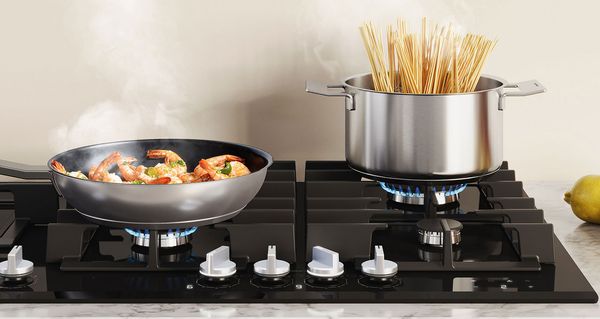 Une casserole contenant des crevettes et une grande marmite contenant des pâtes sont placées sur la plaque de cuisson au gaz.