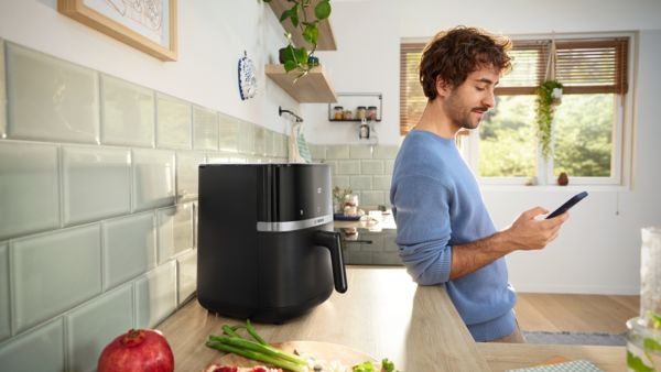 Um homem encosta-se a um balcão de cozinha, olhando para o seu smartphone. Em cima do balcão está uma Air Fryer Serie 4 da Bosch, com uma tábua de corte de madeira perto, exibindo vários legumes frescos, prontos a cozinhar.