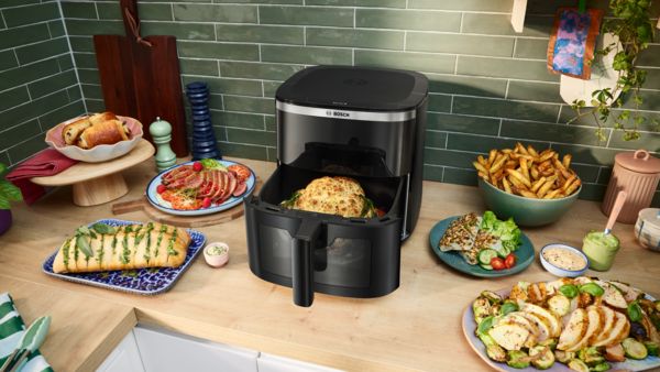 A gaveta aberta de uma Air Fryer Serie 6 da Bosch mostra frango acabado de cozinhar. Ao lado, legumes cozinhados como batatas, couves-de-bruxelas, espargos e beringelas repousam no balcão.