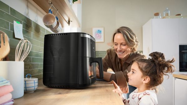 Uma mãe e a sua filha, ambas a observar ansiosamente através da janela de vidro de uma Air Fryer Serie 6 da Bosch, enquanto os alimentos cozinham no interior.