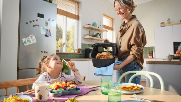 Uma mãe sorri enquanto mostra à filha a gaveta da Air Fryer da Bosch com frango acabado de cozinhar. A filha, sentada à mesa da cozinha com pratos de batatas fritas e legumes, sorri de volta enquanto segura uma batata frita.