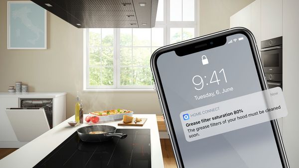 Ένα χέρι κρατάει ένα smartphone, στην οθόνη του οποίου προβάλλονται οι ρυθμίσεις του απορροφητήρα. Στο φόντο, ένα τηγάνι είναι ακουμπισμένο πάνω στη βάση εστιών, ολοκληρώνοντας την ατμόσφαιρα της κουζίνας.