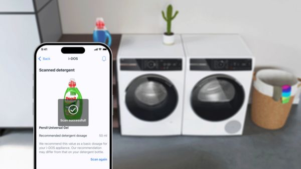 Ένα χέρι που κρατάει ένα smartphone όπου προβάλλονται έξυπνα στοιχεία ελέγχου για δύο πλυντήρια ρούχων, που είναι τοποθετημένα στο φόντο της εικόνας.