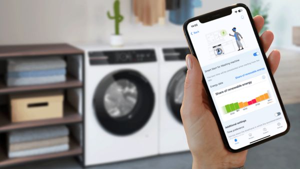 Ένα χέρι κρατάει ένα smartphone που δείχνει ρυθμίσεις συσκευής μπροστά από ένα πλυντήριο και στεγνωτήριο σε ένα φωτεινό δωμάτιο.