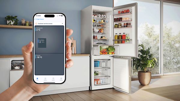 Ένα smartphone όπου προβάλλονται τα περιεχόμενα ενός ψυγειοκαταψύκτη μέσω της εφαρμογής Home Connect. Στο φόντο, φαίνεται στην κουζίνα ο ανοιχτός ψυγειοκαταψύκτης, ο οποίος προσφέρει μια ματιά στην άνετη και σύγχρονη διαχείριση τροφίμων.