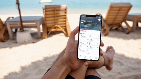 Ένα smartphone που παρουσιάζει την αρχική οθόνη του Home Connect με φόντο ένα έπιπλο κουζίνας και έναν ανοιχτό ψυγειοκαταψύκτη.
