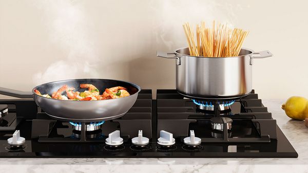 Une casserole contenant des crevettes et une grande marmite contenant des pâtes sont placées sur la plaque de cuisson au gaz.