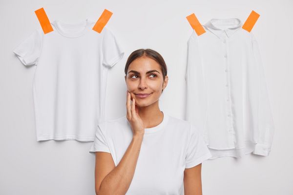 Kobieta w białym T-shircie na tle białych bluzek zastanawia się, jak o nie dbać.