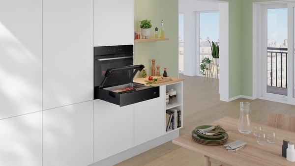 O bucătărie modernă, luminoasă şi spaţioasă, cu un cuptor încorporabil negru şi un sertar termic deschis.