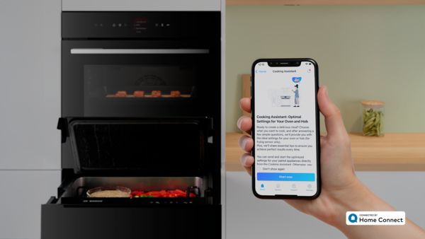 O persoană ţine în mână un smartphone pe care se observă o aplicaţie de asistare la preparare, iar în fundal se observă un cuptor deschis.