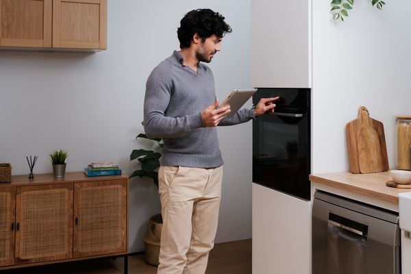 En mand i et moderne køkken bruger en tablet til at betjene en sort, indbygget ovn fra Bosch.