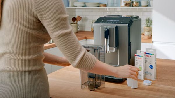 Une personne en manches longues beiges débouche un réservoir d'eau pour une machine à café Bosch, sur une table en bois clair dans une cuisine lumineuse, avec des cartons de produits Bosch à côté.