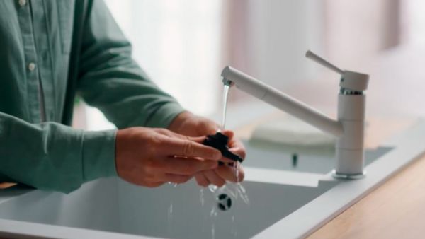 Une personne portant une chemise verte lave un élément (probablement un filtre ou un accessoire) sous un robinet qui coule dans un évier moderne. L'eau s'écoule et goutte des mains de la personne, tandis que l'évier est illuminé par la lumière naturelle entrant par la fenêtre.
